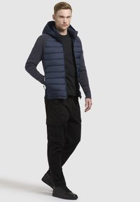 Gilet rembourré pour homme avec un devant matelassé bleu marine, des manches grises, une capuche ajustable et un pantalon cargo noir. Les détails accentués comprennent des poches zippées.