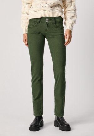 Slim fit olijfgroene broek met dubbele knopen, zijzakken, gedragen met een crèmekleurige gebreide trui en zwarte stevige laarzen.