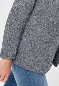 Blazer gris texturizado con un solo bolsillo en el pecho. Presenta mangas ajustadas y una forma estructurada, diseñado en un tejido tejido.