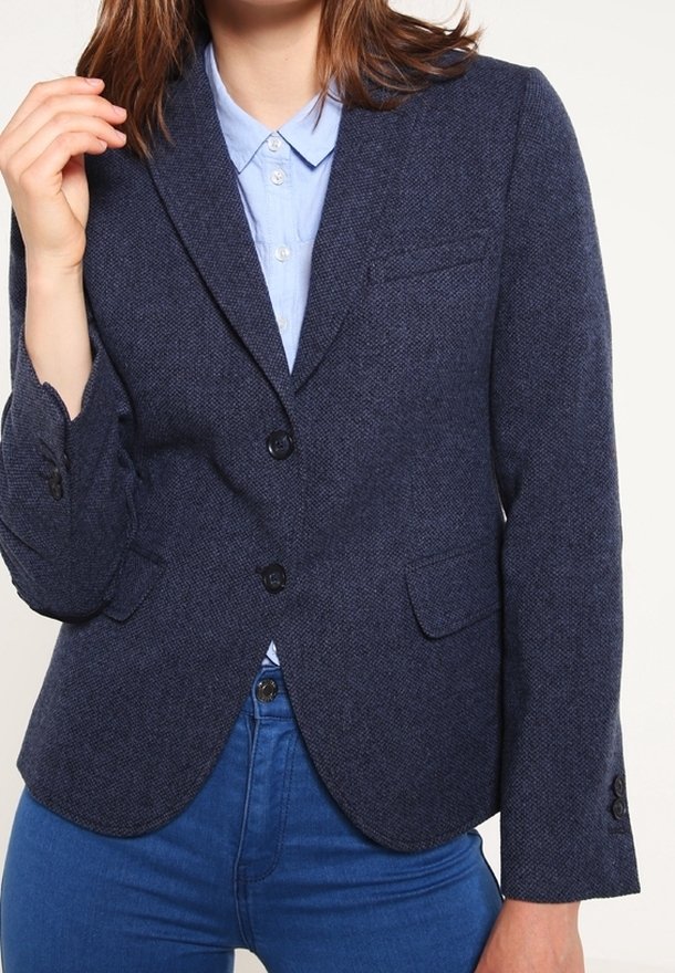 Femme portant un blazer bleu foncé sur une chemise bleu clair boutonnée et un jean bleu, une main levée près de son visage.