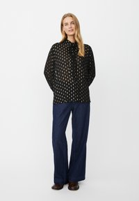 Schwarze durchsichtige Bluse mit goldenen gepunkteten Mustern, Blusenkrawatte, lange Ärmel, kombiniert mit hochgeschnittenen, weiten Jeans in dunkler Farbe.