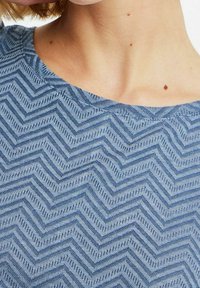 Top azul con textura que presenta un patrón de zigzag, con un escote liso y un brillo sutil. El material parece ligero y transpirable.