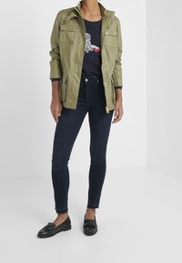 Chaqueta ligera verde oliva con dos bolsillos frontales, camisa azul marino con un diseño gráfico, jeans ajustados oscuros y mocasines negros.