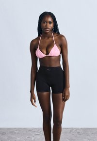Ružový trojuholníkový bikinový top s tenkými ramienkami v kombinácii s čiernymi vysokým pásom krátkymi nohavicami, ktoré majú hladkú textúru a priliehavý strih.