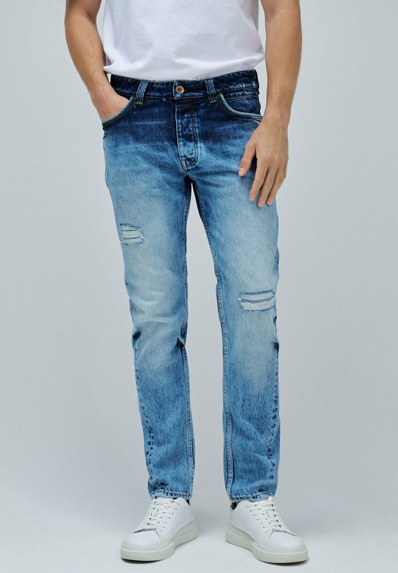 Jeans de mezclilla azul claro con un lavado desvanecido, que presentan dos desgastes en las rodillas y un corte recto clásico, combinados con zapatillas blancas.