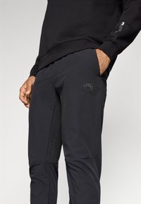 Black athletic pants with a tapered design, featuring a textured panel and embroidered logo. Made from lightweight, flexible material.