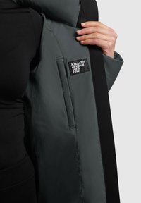 Abrigo acolchado gris con paneles laterales negros, que presenta una etiqueta "Khujo Original for a few" en el interior, mostrado con una mano sosteniendo la tela.