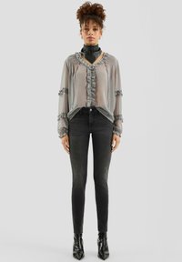 Przezroczysta szara bluzka z falbanami, zestawiona z dopasowanymi czarnymi jeansami i czarnymi, szpiczastymi botkami na kostkę. Modelka stoi na gładkim tle.