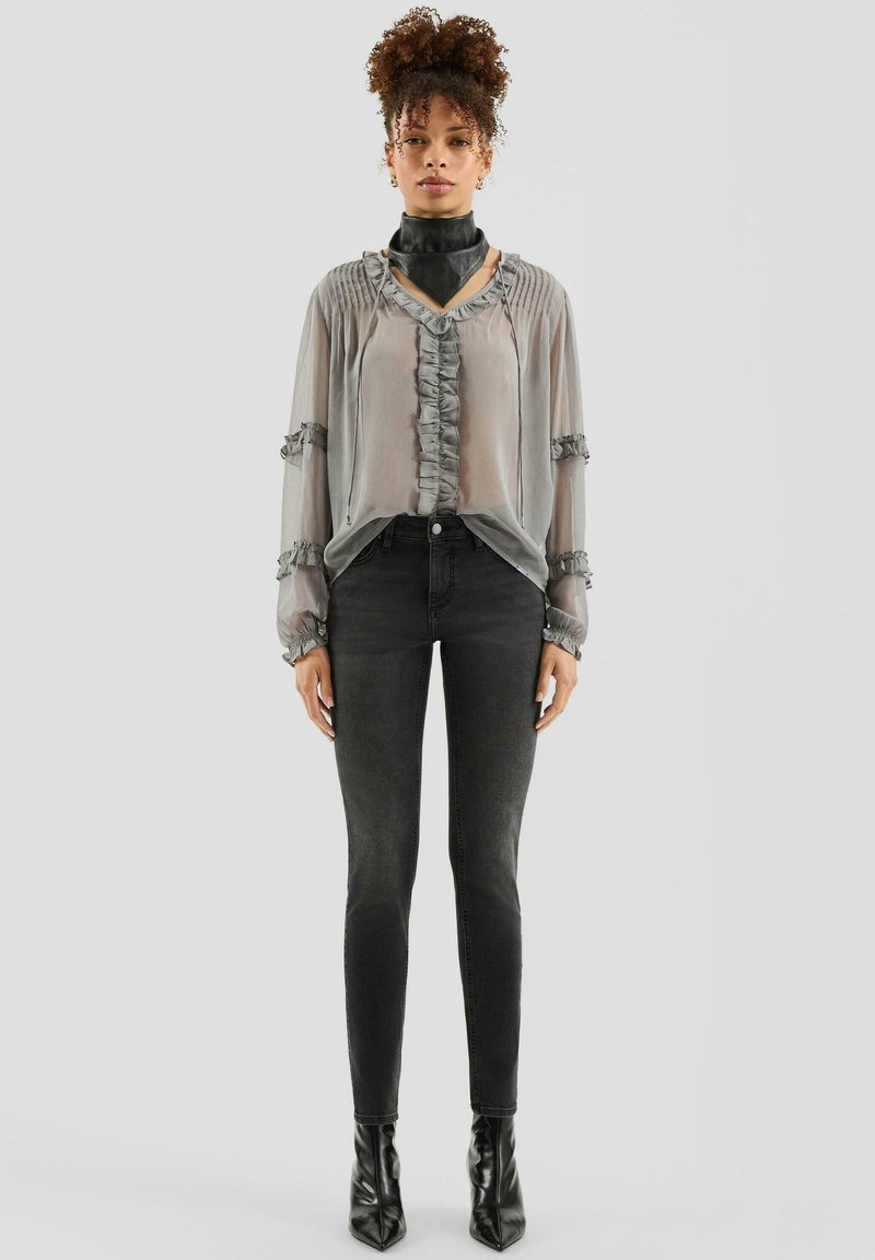 Przezroczysta szara bluzka z falbanami, zestawiona z dopasowanymi czarnymi jeansami i czarnymi, szpiczastymi botkami na kostkę. Modelka stoi na gładkim tle.