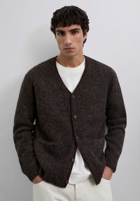 Joven con cabello rizado oscuro, vistiendo un cárdigan marrón oscuro abotonado sobre una camisa blanca y pantalones de color claro, con las manos en los bolsillos.