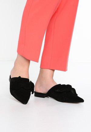 Mules de gamuza negra con punta en forma de pico, que presentan un gran lazo decorativo en la parte delantera y un tacón bajo. Combinados con pantalones cortos color coral.