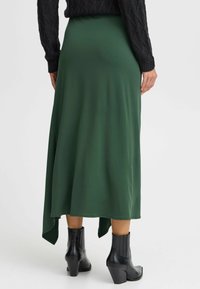 Grön, flödande maxikjol med asymmetrisk fåll, tillverkad av mjukt tyg, ihop med svarta klackade ankelstövlar. Texturerad stickad tröja ovanpå.