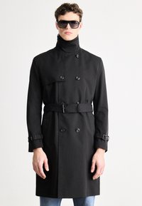 Mustast trentšmantlist, millel on topeltsuunalised nööbid, vöökohal rihm, kõrge krae ja reguleeritavad varrukapikendused. Modell kannab päikeseprille ja seisab lihtsa taustaga keskkonnas.