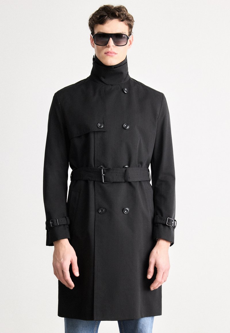 Mustast trentšmantlist, millel on topeltsuunalised nööbid, vöökohal rihm, kõrge krae ja reguleeritavad varrukapikendused. Modell kannab päikeseprille ja seisab lihtsa taustaga keskkonnas.