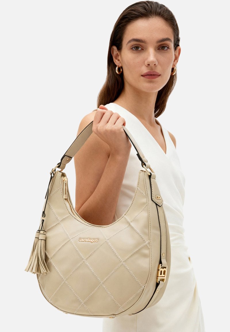 Borsa hobo in pelle trapuntata beige con accenti dorati, nappina e tracolla removibile. Presenta un design spazioso e una texture liscia.