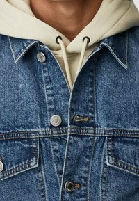 Persona con una sudadera con capucha de color blanco roto debajo de una chaqueta vaquera azul con botones metálicos visibles y bolsillos delanteros en el pecho.