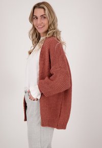 Roestkleurige gebreide cardigan met een gestructureerd patroon, open voorkant en een losse fit. Gecombineerd met een witte blouse en gestreepte broek.