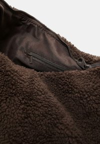Hnedá fleecová bunda s hladkým tmavým podšívkovým materiálom a zipsovým vreckom. Textúra je mäkká a nadýchaná. Viditeľný detail zipsu.