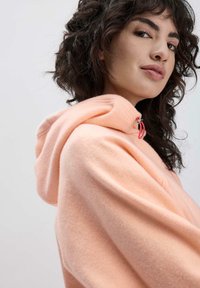 Pfirsichfarbener Kapuzenpullover mit weicher Textur, ausgestattet mit einer Zugkordel und einer abgerundeten Kapuze, getragen von einem Modell vor einem hellen Hintergrund.