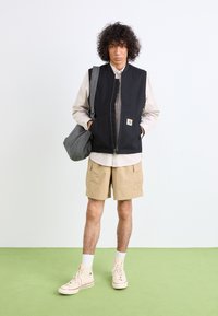 Schwarze Zip-Weste mit Seitentaschen, beiges Hemd, khakifarbene Cargoshorts. Trägt beiges High-Top-Sneaker mit weißen Akzenten. Grauer gemusterter Beutel.