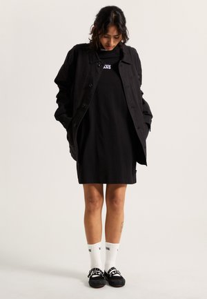 Giubbotto di jeans nero, abito t-shirt nero, calzini bianchi a coste, e scarpe nere con dettagli circolari. Design minimalista, vestibilità casual.