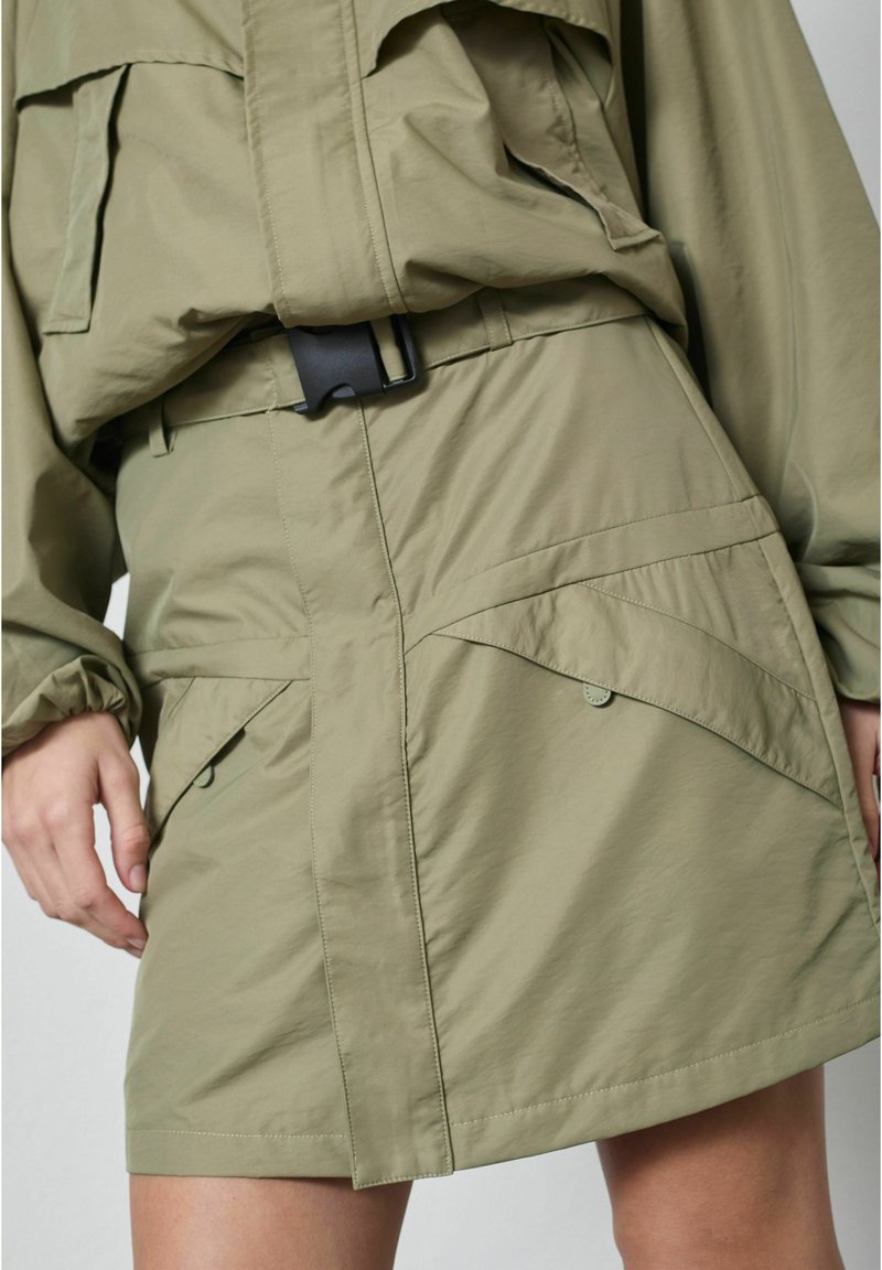 Olivovo zelená sukňa s chlopňovými vreckami a čiernym opaskom na pracku, nosená s voľnou zodpovedajúcou bundou, ukazujúca ruky a hornú časť stehien.