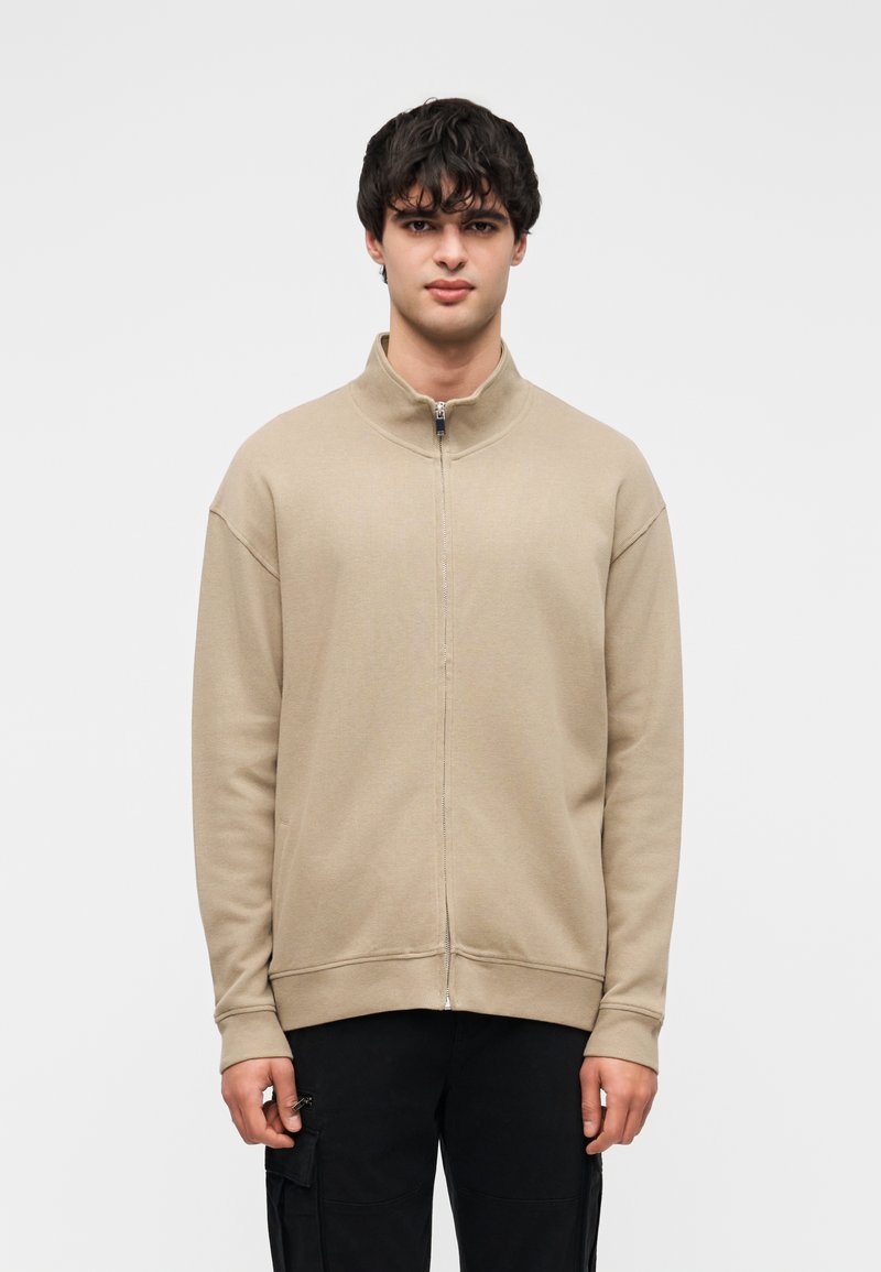 Μπεζ φούτερ με φερμουάρ, ψηλό γιακά, ριμπ μανσέτες και ίσια τελική ράχη. Με λεία υφή και άνετη εφαρμογή.