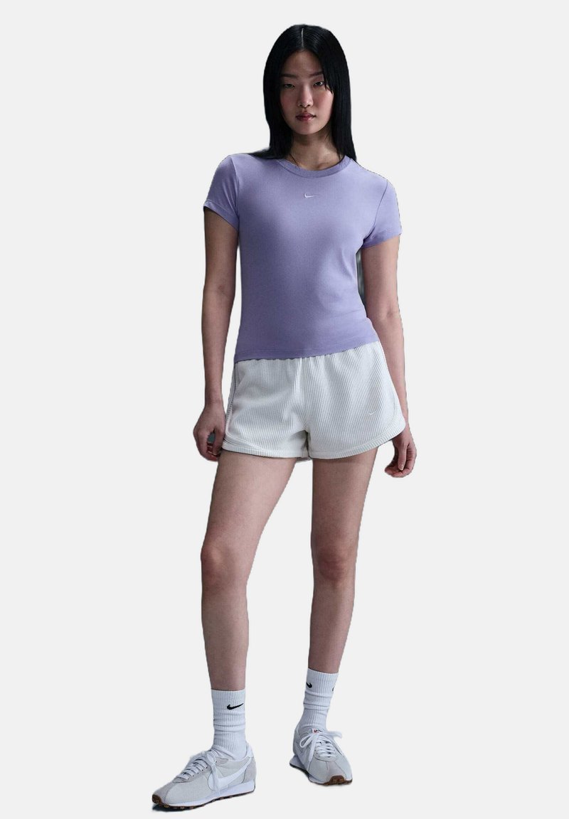Lila sportieve top met korte mouwen, gecombineerd met witte geribbelde shorts. Het model draagt witte sneakers en crew socks met een klein Nike-logo.