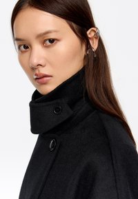 Joven con cabello largo y oscuro, vistiendo un abrigo negro de cuello alto y múltiples aros de plata en las orejas, mirando hacia la cámara.