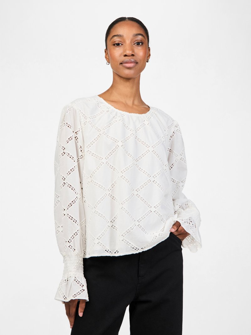 White blouse with diamond cut-out pattern, round neckline, and long sleeves with flared cuffs. Fabric is lightweight and textured.
