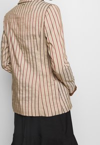 Blazer a righe in tessuto beige con strisce verticali rosse e nere. Presenta un colletto classico e una fessura sul retro. Indossato sopra una gonna nera.