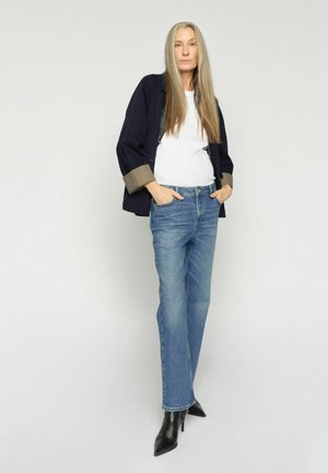 Žena s dlhými rovnými vlasmi, oblečená v námorníckej bunde, bielom tričku, modrých džínsoch a čiernych špicatých vysokých čižmách, stojaca so založenými rukami v kapsách.