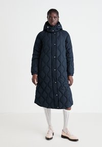 Marineblauwe gewatteerde lange jas met een aansluitende capuchon, drukknoopsluiting en ribgebreide manchetten. Model draagt witte knie-sokken en roze veterschoenen.