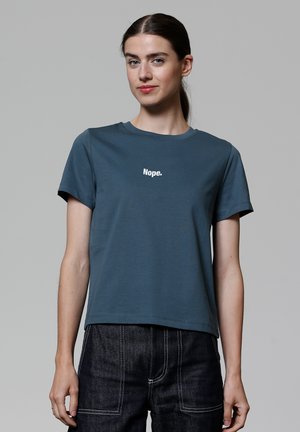 T-shirt en coton teal à manches courtes, col rond, arborant le mot "Nope." en texte blanc sur la poitrine. Porté avec un jean foncé.
