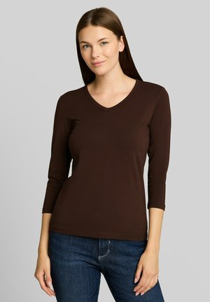 Vrouw met steil bruin haar die een donkerbruin V-hals shirt met lange mouwen en een blauwe spijkerbroek draagt en staat tegen een effen lichtgrijze achtergrond.