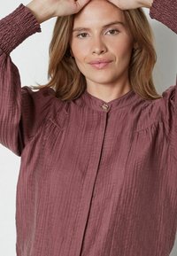 Szilva színű, texturált blúz magas gallérral, plisszírozott részletekkel és egy kis kör alakú gombbal a nyakán. A puffos ujjú rész a csuklónál ráncolódik.