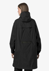 Veste imperméable noire avec capuche, design oversized, poignets élastiques, poche arrière avec fermeture éclair et texture lisse et légère.