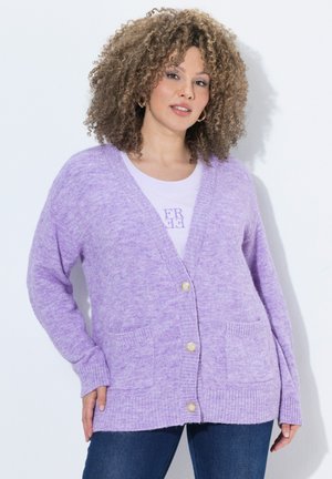 Kvinde iført en lyse lilla knap-cardigan over en hvid skjorte med tekst, kombineret med mørkeblå jeans, stående mod en hvid baggrund.
