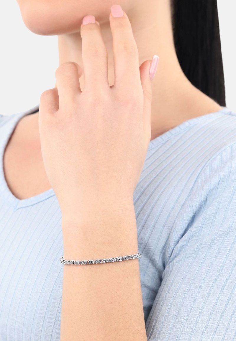 Silverfärgad armband med intrikata bladmönster, slät yta och flexibel design, bärs på ett ljust skinns handled. Ljusblå topp.