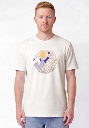 Man met kort rood haar die een wit T-shirt draagt met een cirkelvormig abstract ontwerp in paars en geel, staand tegen een effen witte achtergrond.