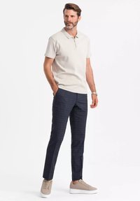 Beige polo tričko s krátkým rukávem a zipem, spárované s tmavě kostkovanými kalhotami a světle béžovými teniskami. Jednoduchý, moderní střih.