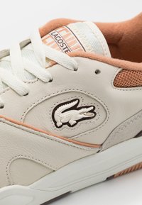 Lacoste sneakers in cream leather with peach accents. Features textured mesh panels, a signature crocodile logo, and flat laces.