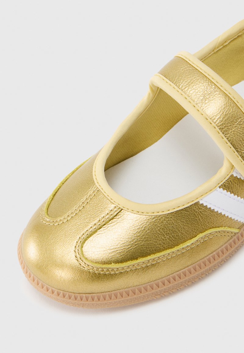 Gold metallic Mary Jane shoe with a round toe, strap across the instep, white accent stripe, and textured rubber sole.