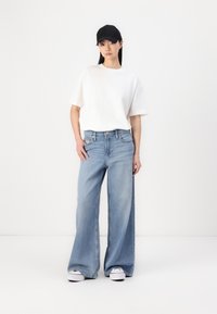 Jeans larghi di colore blu chiaro abbinati a una maglietta bianca e un cappellino nero. I jeans presentano uno stile classico a cinque tasche e una vestibilità rilassata.