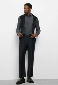 Dunkelgraues Langarmhemd mit dezentem Muster, schwarze Strickweste und maßgeschneiderte dunkle Hose; schwarze Schuhe mit strukturierter Oberfläche.