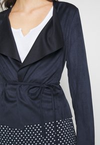 Blazer blu navy con colletto nero a contrasto, cintura in vita e texture liscia, indossato sopra una camicia bianca con scollo a V e una gonna a pois.