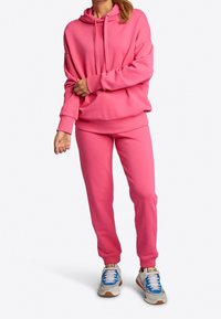 Różowa bluza z kapturem i sznurkiem, luźny krój; dopasowane różowe dresy z elastycznymi mankietami. Noszone z wielokolorowymi trampkami. Gładka faktura.