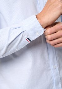 Svetle modrá pruhovaná košile na knoflíky s bílými knoflíky, s klasickým límcem a malou emblémem v červené, bílé a modré barvě na manžetě.