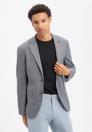 Young man wearing a grey blazer over a black shirt and light blue pants, adjusting the blazer with one hand.