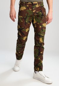 Spodnie w kamuflażu w kolorze brązowym, zielonym i beżowym o dopasowanym kroju. Noszone z białymi sneakersami, z kieszeniami i wytrzymałą fakturą materiału.